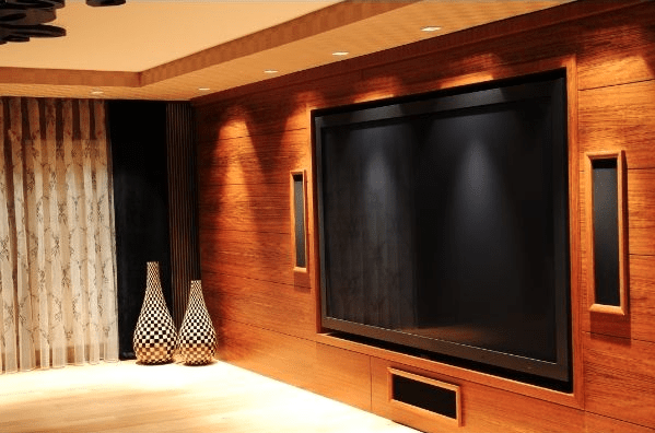 Modern living room with a large, wall-mounted flat screen TV framed by wood paneling, two patterned floor vases on the left, and ceiling spotlights illuminating the space.